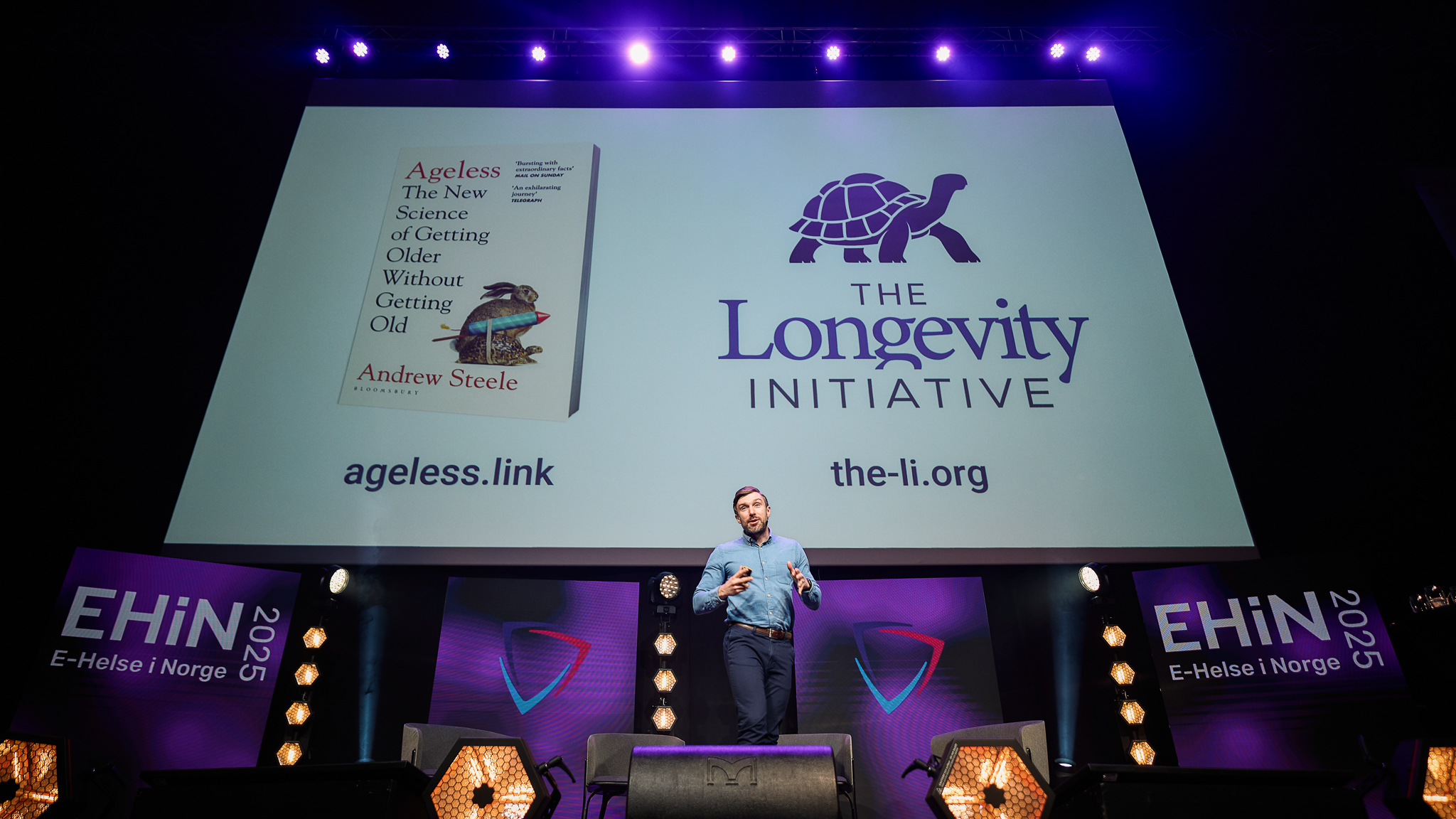 Andrew Steele stood on a purple-lit stage with his book, Ageless, and The Longevity Initiative’s logo on a huge slide behind him